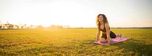 Person smiles while in a yoga pose on a mat, lit by the morning sun, in a field