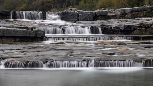 An impressive, multi-levelled waterfall flows over stones in a tranquil natural setting, Val