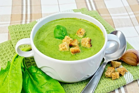 Green soup puree in a white bowl with croutons and spinach leaves, spoon on a napkin on the