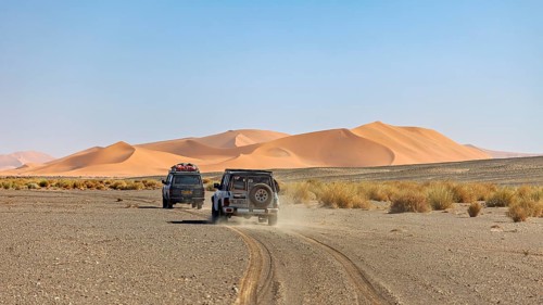 Off-road vehicles drive on a dusty road in front of large sand dunes, With off-road vehicles