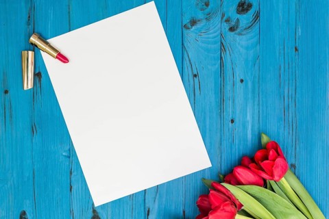 Red tulips, lipstick and a white sheet of paper for your greetings on the background of blue wooden