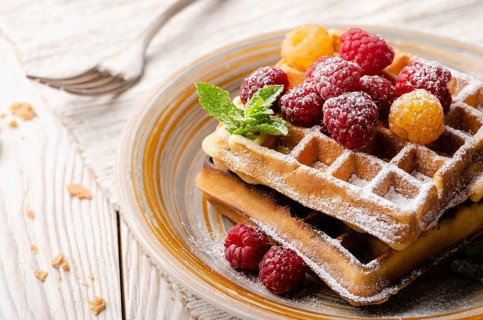 Belgian waffles served with raspberries and mint leaf dusted with powdered sugar on white wooden