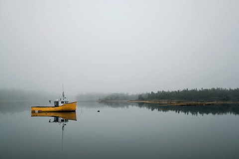 A commercial fishing boat is reflected in the still calm water of a protected bay as light fog