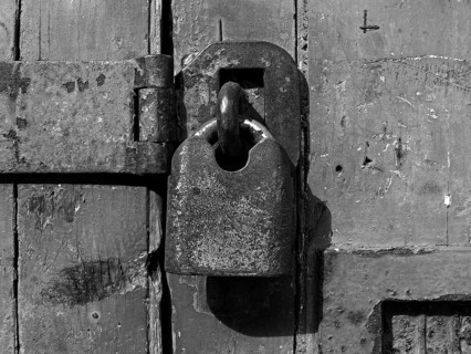 A monochrome close up of an old rusty closed padlock and hasp on a distressed wooden plank door