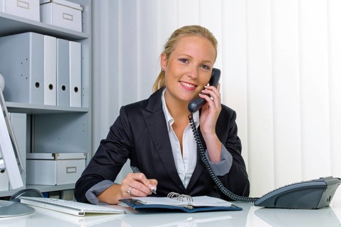 A friendly woman talks on the phone at her desk in the office and notes appointments in her