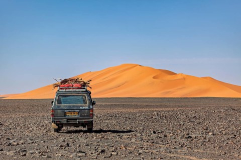 Off-road vehicle leads through a rocky plain in front of large sand dunes under a blue sky, By