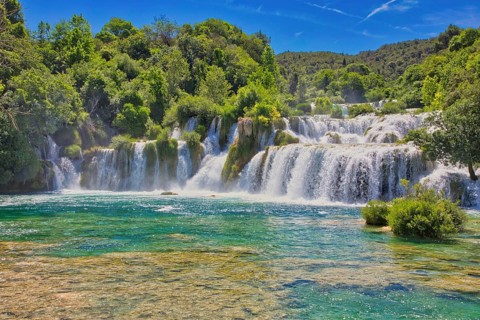 Waterfalls flow into an emerald green river, surrounded by dense forest and a bright blue sky, Krka