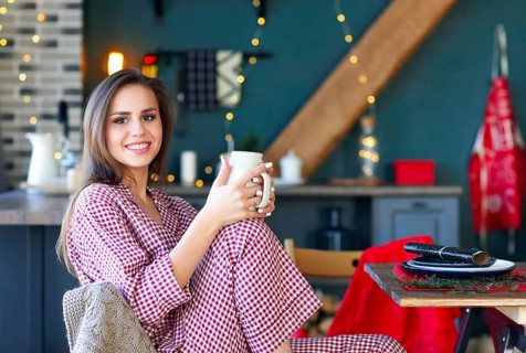 Happy young woman in plaid pajama holding jar smiling at camera in kitchen decorated for Christmas
