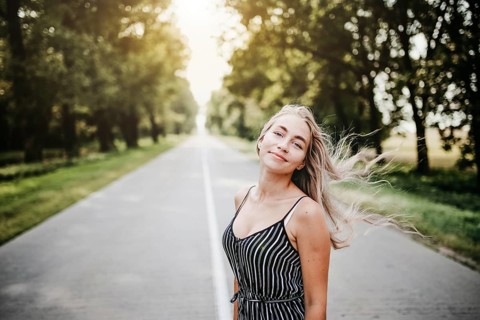 Woman standing on a road surrounded by trees, sunlight filtering through, creating a relaxed summer