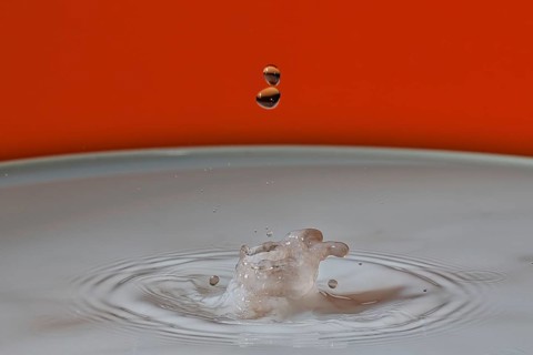 A beige water droplet hits the water surface and creates a small splash on an orange background,