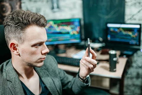 Man examining a smartphone in an office setting with trading monitors behind