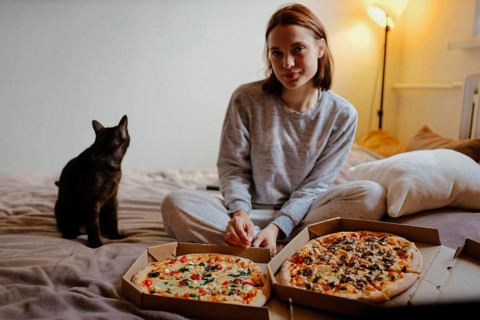 Woman sitting on the bed with pizza boxes and a black cat in a nighttime cozy setting