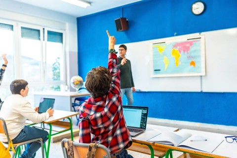 Elementary school student raising hand to answer a question in geography class, with teacher and