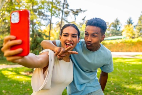 Multi-ethnic couple taking selfie gesturing victory and blowing a kiss in a park