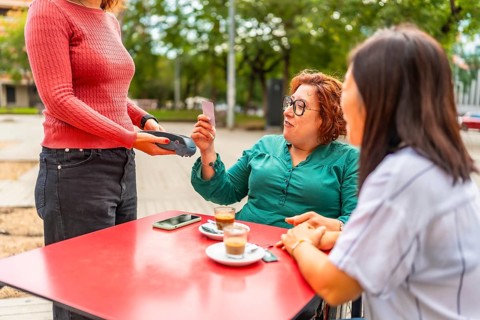Woman with disability in wheelchair and friend paying with contactless in an outdoor cafeteria