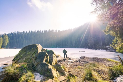 Man standing on rocks in front of a frozen lake, surrounded by forest, sunlight illuminates the
