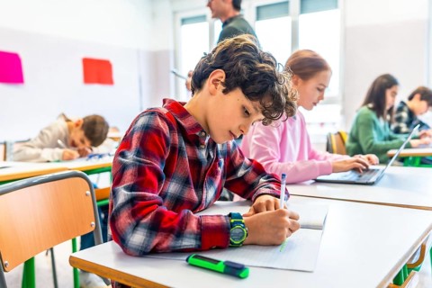 Elementary school students are diligently working at their desks, some writing in notebooks and