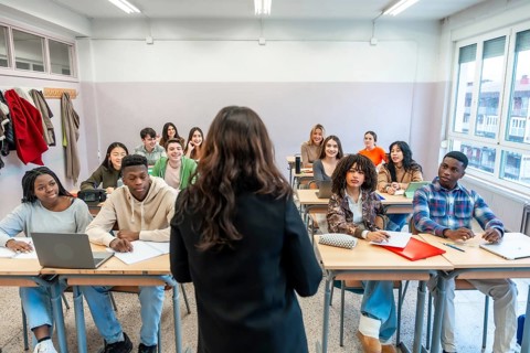Female professor teaching a lesson to a multi ethnic group of students sitting at desks in a