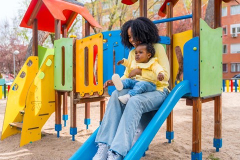Happy African mother with baby girl on lap sliding in a colorful playground