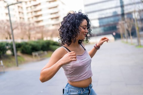 Curly haired hispanic woman joyfully dancing in an urban setting, embracing the sunny day with