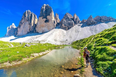 Tourist hiking along a scenic trail by a crystal clear alpine lake, with the stunning tre cime di