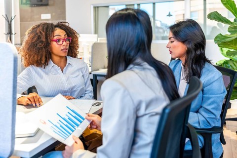 Three businesswomen are discussing over financial charts sitting at a desk in a modern coworking