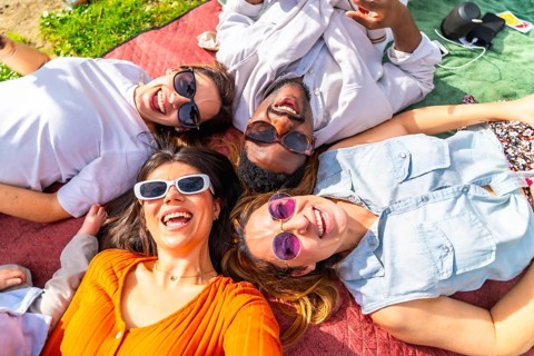 Four cheerful young adults lying together on blanket, enjoying sunny day picnic in park, listening