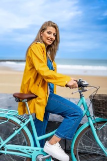 Young woman with yellow raincoat sitting on a turquoise vintage bicycle by the beach, enjoying a