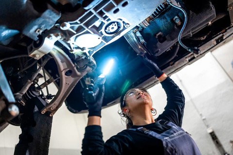 Female auto mechanic inspecting the underside of a car in a repair shop, using a flashlight to