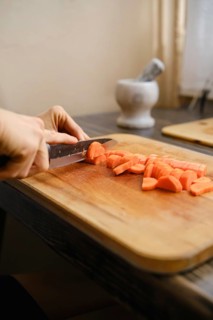 A person skillfully slices fresh carrots into bite-sized pieces on a wooden cutting board. Sunlight