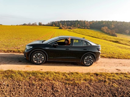 A driver sits in a black car on a dirt track, surrounded by green meadows, electric car VW ID5,