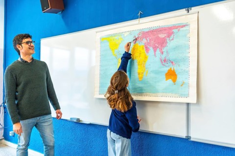 Smiling teacher watching elementary school student pointing at a world map during geography class