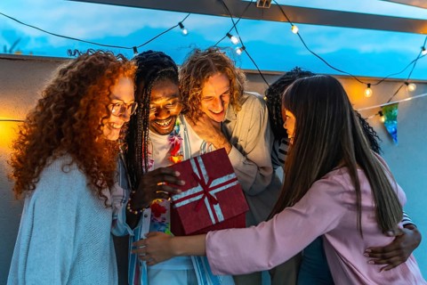 African man opening birthday present embraced by his multiracial friends during rooftop party