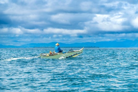 Traditional outrigger boat sailing on turquoise water with a fisherman wearing a straw hat, near