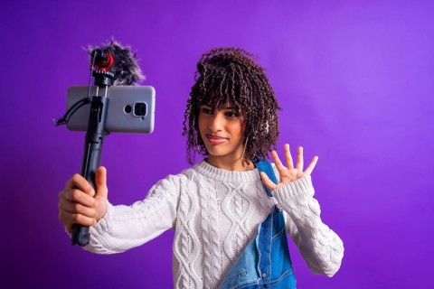 Young influencer waving while recording a video blog using a smartphone with external microphone on