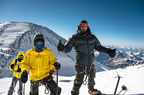 A middle-aged climber in a down jacket and harness shows a thumb up next to his friends on the way