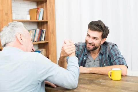 Aged man young happy guy with hands clasped arm wrestling challenge table room