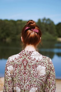 rear view of anonymous young woman with floral dress and red hair bun standing by lake in solitude