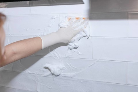 Gloved hand spreading foam cleaner on white tiled wall