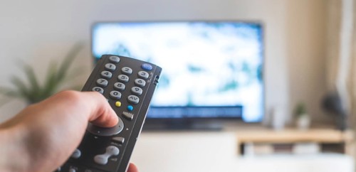 Holding a TV remote control in the hand, foreground, tv in the blurry background. Streaming