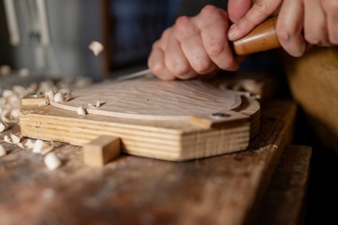 Luthier using gouge and mallet, shaping wood for a violin in his workshop, surrounded by wood