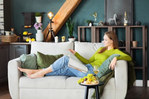 Side view of young female reading interesting magazine while lying on comfortable couch near table