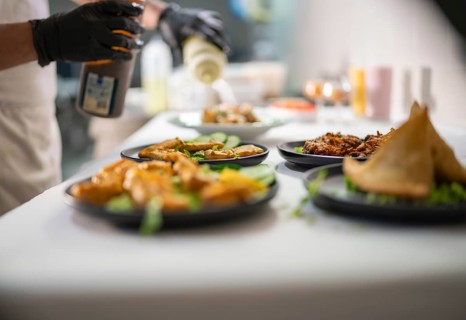 Chef wearing black gloves is pouring a sauce from a bottle on several plates of indian food,