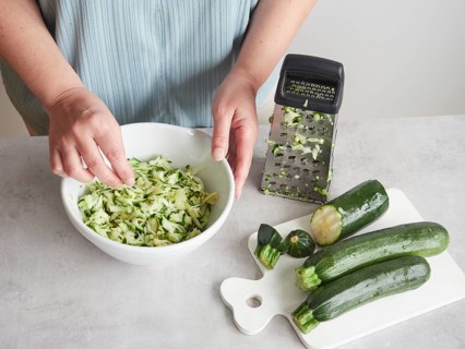 Woman with her hands in a bowl of grated zucchini, other fresh zucchini on wooden board and