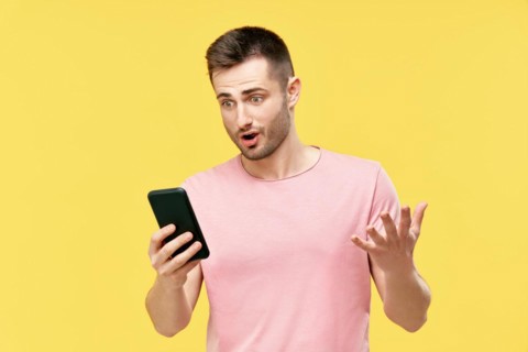 Shocked funny millennial man using mobile phone holding it in hand isolated over yellow background