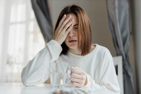 Woman holding her forehead, looking unwell, with a glass of water and curtains in the background