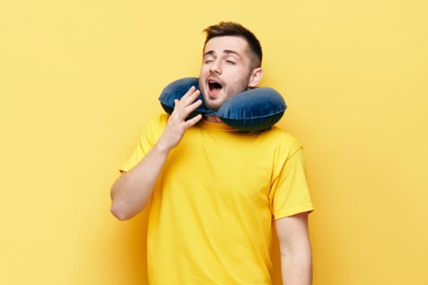Tired young man yawning with travel pillow on yellow background. tourism, rest concept