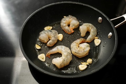Raw peeled shrimps and garlic are sauteed in a cooking pan on the black stove, preparation for a