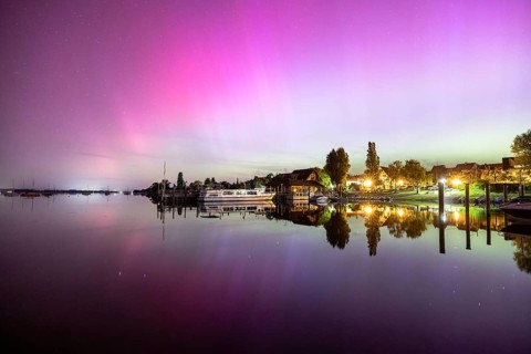 Colourful northern lights reflected in a quiet night lake next to illuminated buildings, harbour,