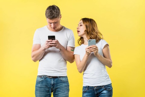 Suspicious girlfriend spying his boyfriend texting mobile phone against yellow backdrop